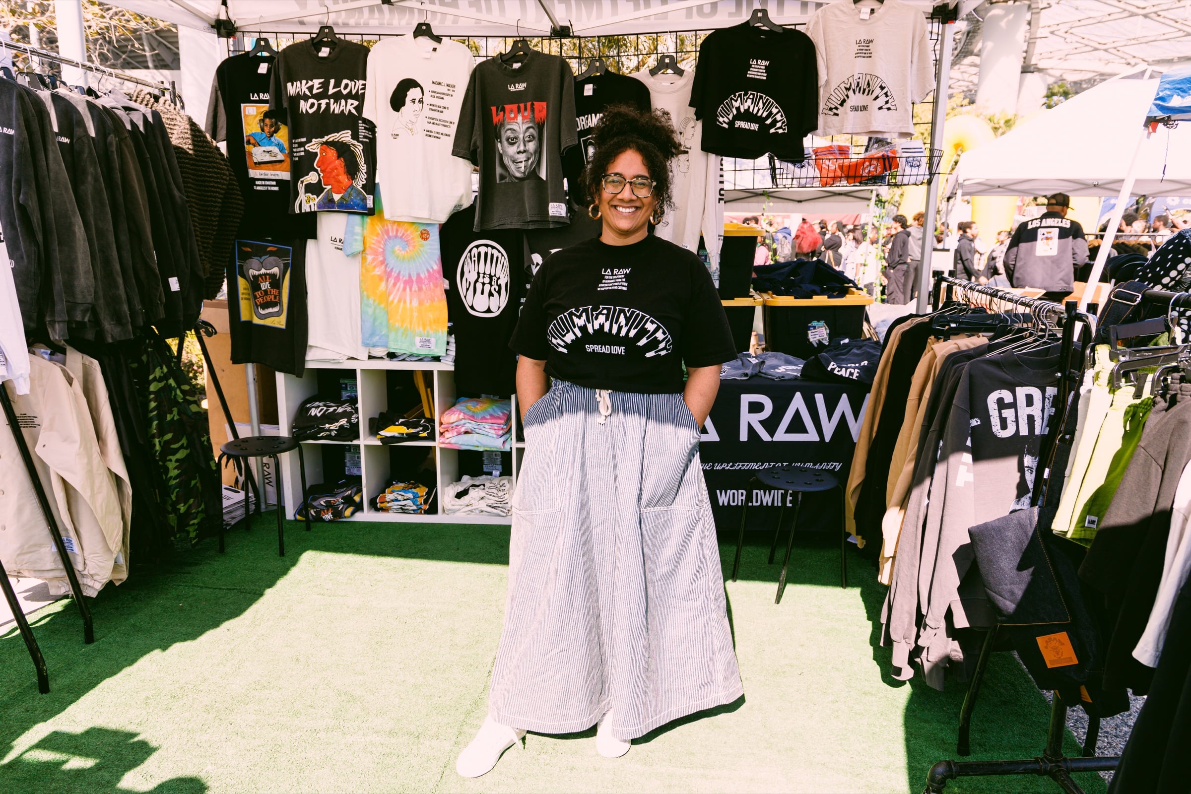 Smiling vendor standing among printed T-shirts and clothing at an outdoor booth